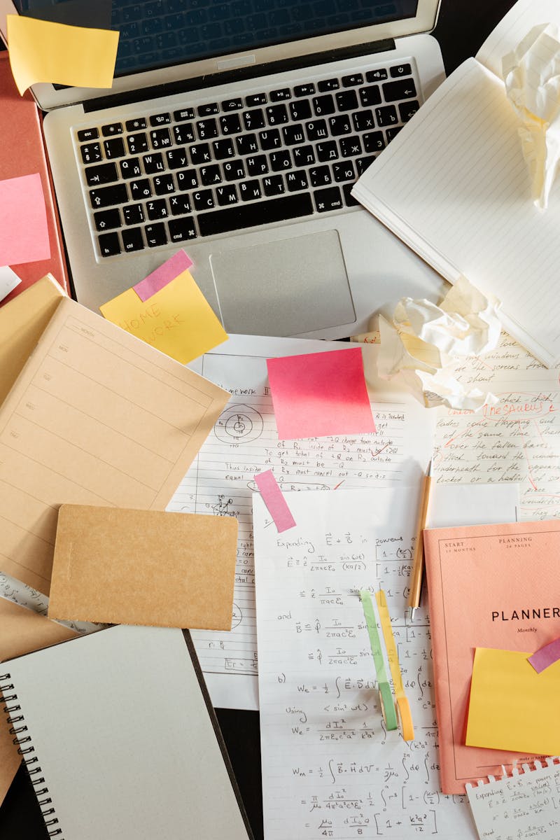 Flatlay of a messy study desk with laptop, notebooks, and sticky notes, showcasing the creative process.